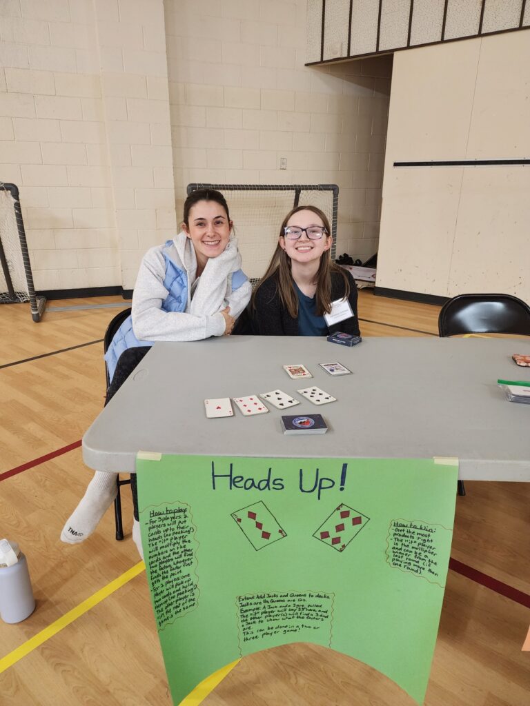 Two people sitting at a table with cards on it and a sign that says "Heads Up!" with game instructions.